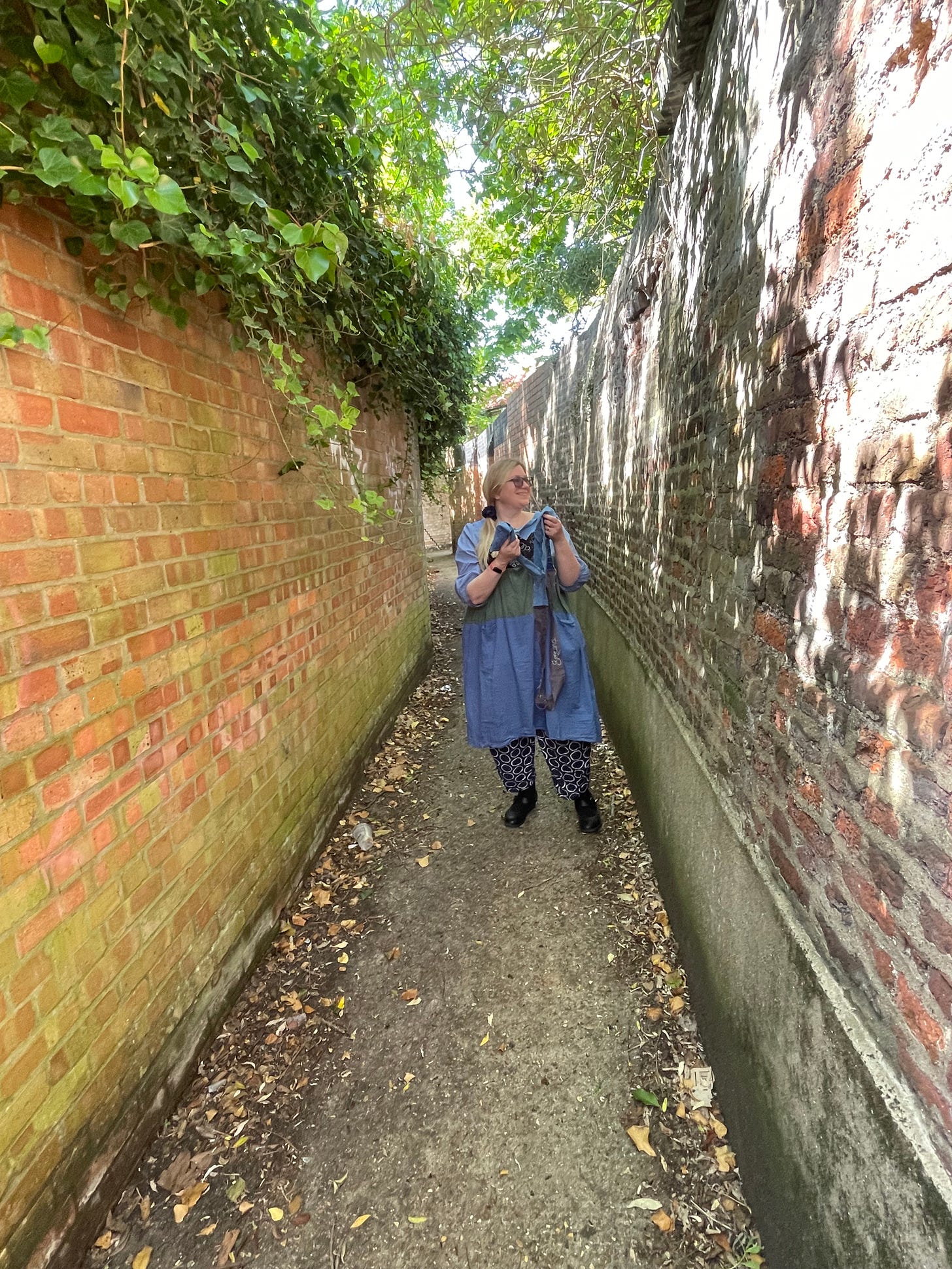 A white woman with blonde hair and a blue dress stands in between two walls. The wall on the left is modern, with ivy on the top. The wall on the right is much older, and cracked. She is holding fabric pieces.