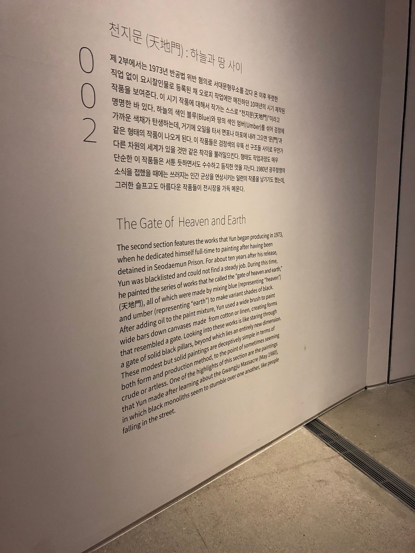 The museum description for The Gate of Heaven and Earth Series. It says, “The second section features the works that Yun began producing in 1973, when he dedicated himself full-time to painting after having been detained in Seodaemun Prison. For about ten years after his release, Yun was blacklisted and could not find a steady job. During this time, he painted the series of works that he called the "gate of heaven and earth," all of which were made by mixing blue (representing "heaven" and umber (representing "earth") to make variant shades of black. After adding oil to the paint mixture, Yun used a wide brush to paint wide bars down canvases made from cotton or linen, creating forms that resembled a gate. Looking into these works is like staring through a gate of solid black pillars, beyond which lies an entirely new dimension. These modest but solid paintings are deceptively simple in terms of both form and production method, to the point of sometimes seeming crude or artless. One of the highlights of this section are the paintings that Yun made after learning about the Gwangju Massacre (May 1980), in which black monoliths seem to stumble over one another, like people falling in the street.