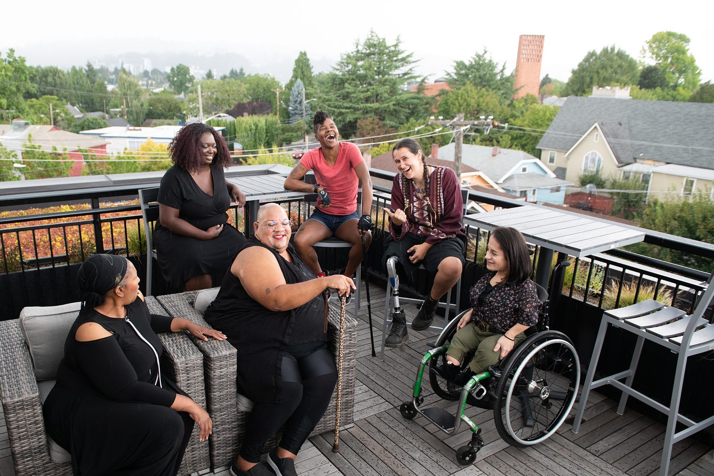 Overhead shot of six disabled people of color at a rooftop deck party. An Indigenous Two-Spirit person with a prosthetic leg smiles directly at the camera and gives a thumbs up while everyone else is engaged in conversation.