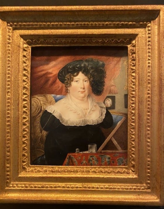 A photograph of a framed watercolour painting. The painting shows a woman looking at the viewer. She has curly dark hair and a flamboyant hat. She has a dark-coloured dress with white lace at the neck. She has no arms and there is a slender paintbrush attached to the sleeve on her right shoulder. In front of her is a table with a patterned tablecloth, a painting slope and a water glass.