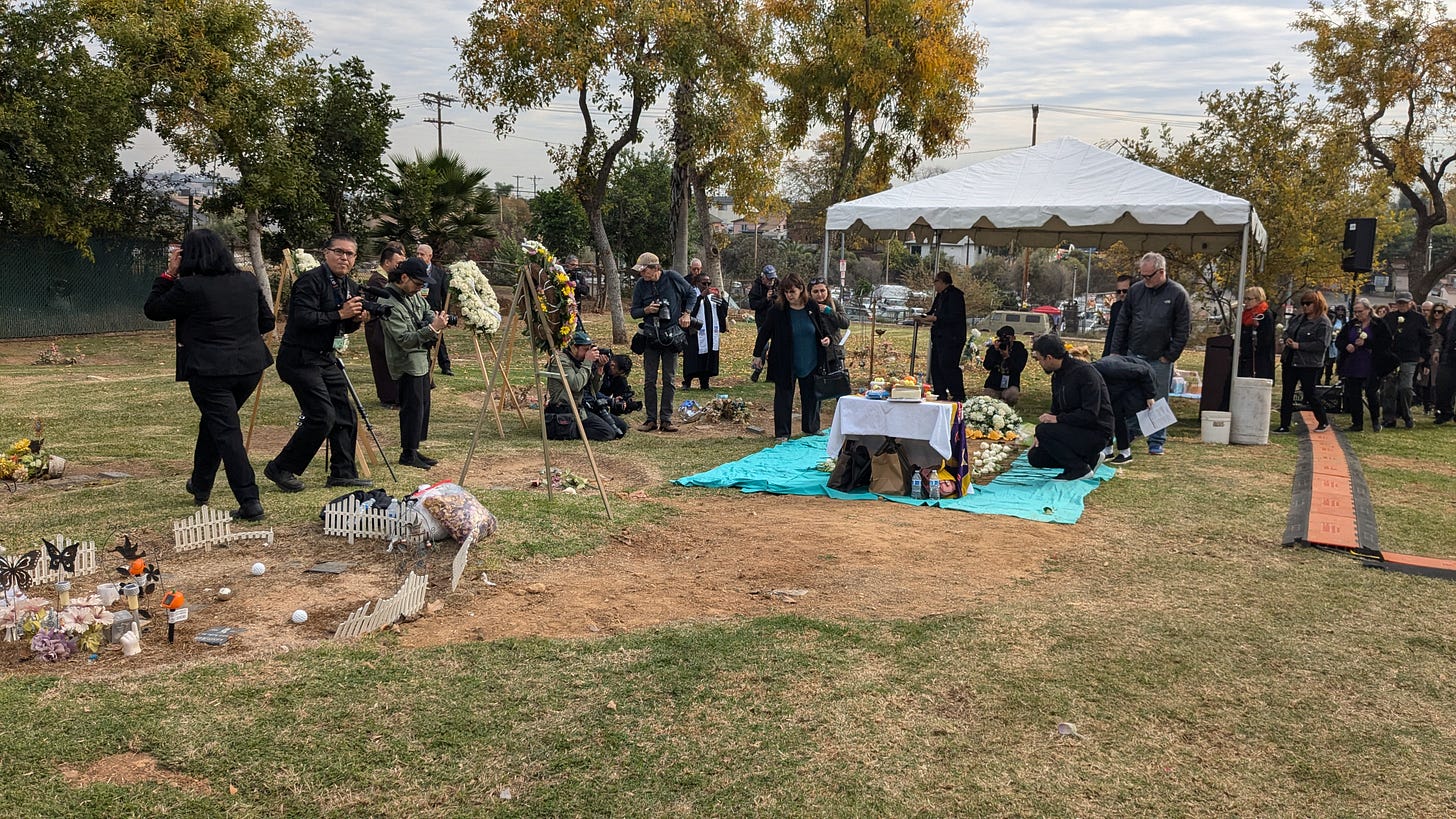 Press photographers watch people line up to place flowers on a mass grave on a grassy hillside