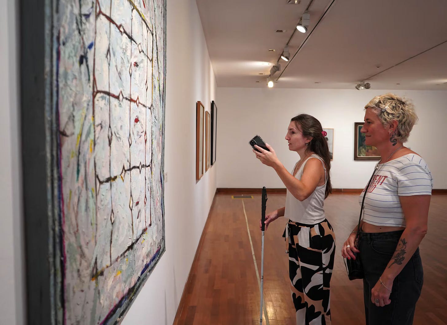 Two women standing before a painting in a museum. One of them is holding a white cane and using her phone.