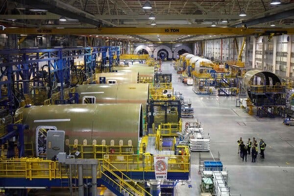 Several plane fuselages on the floor of a factory, with a group of workers standing on the floor.