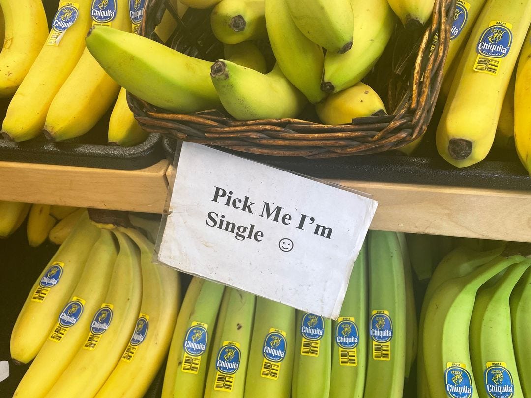 A sign that says "Pick me, I'm single!" hangs between two rows of bananas in a market