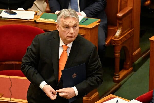 Prime Minister Viktor Orban of Hungary, in a dark suit and orange tie, standing in Parliament amid rows of desks.
