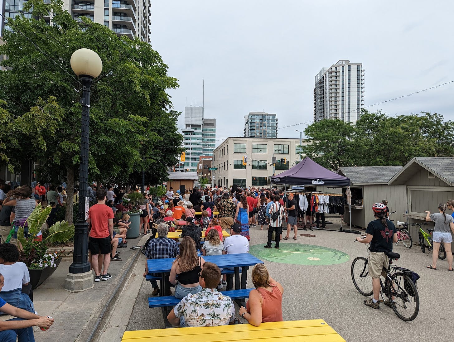 A street with many pedestrians and cyclists and no cars, street trees and tables on one side, pop-up shops on the side with the city skyline in the distance.