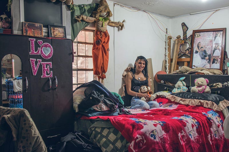 Messy bedroom. Lady sitting on the bed. Red covers. Toys on the bed. Large photo at the head of the bed.