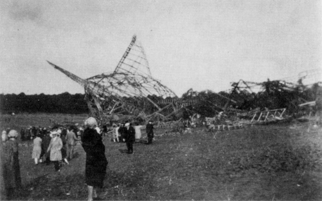 Photograph showing the wreckage of the airship R101 in a field in France with onlookers looking at the charred frame of the airship.