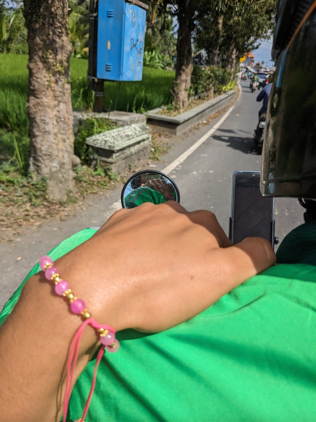 Nathalie holds onto the motorbike driver's shoulder. Her pink and gold bracelet is visible on her wrist. She snaps a photo of herself to show her reflection om the motorbike's rearview mirror