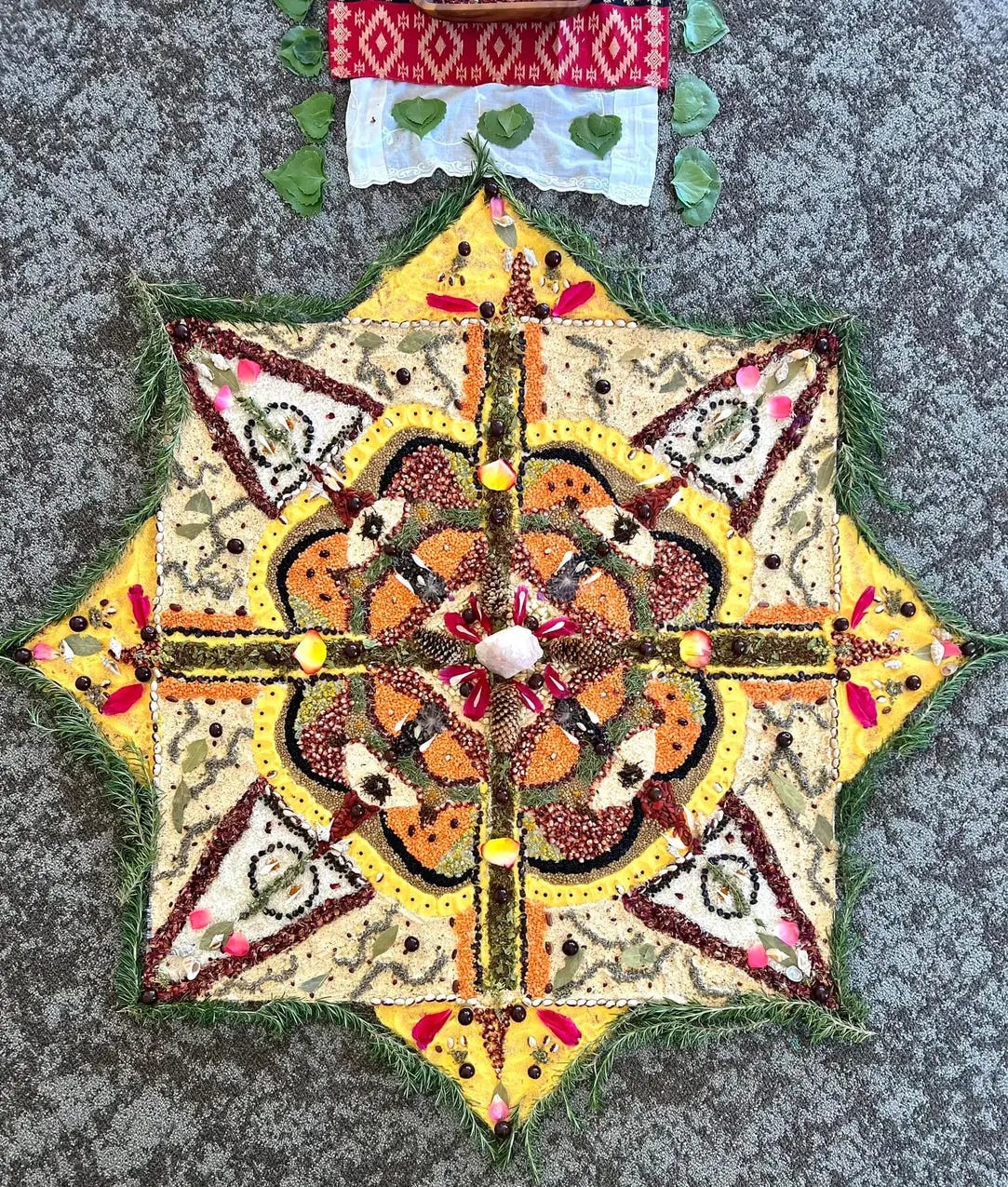 An approximately 3-foot wide, 8-pointed mandala sort of pattern on the floor. Each quadrant is symmetrical with a central flower pattern. Made from plant materials, it is multi-colored in earthy hues of yellow, white, red, orange, black and green. It is outlined in sprigs of rosemary.