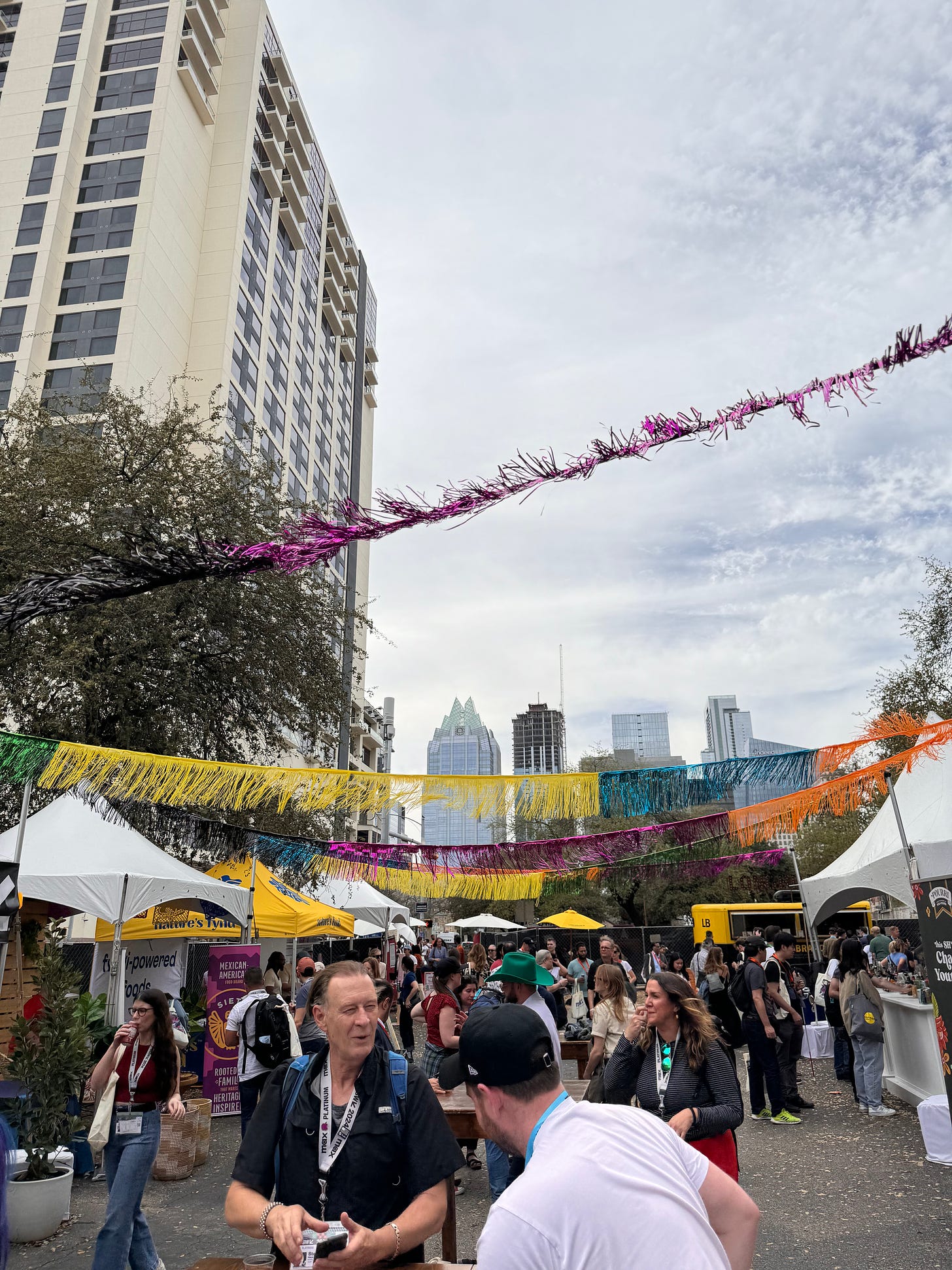 Photo from outdoors SXSW registrant lounge, with colorful streamers and the Austin skyline in the background. Various people mill around and visit the sponsor tents for food/drink.
