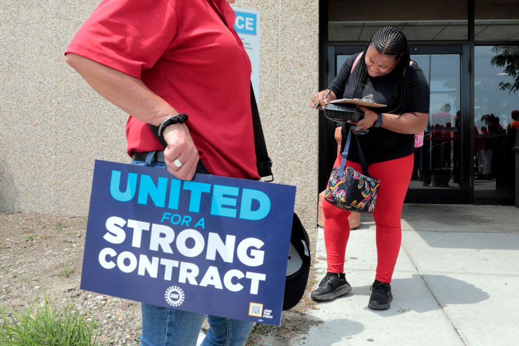 side profile of someone wearing a red shirt and carrying a dark blue sign with light blue and white text that reads "UNITED for a STRONG CONTRACT" with the UAW logo. a Black woman stands off to the side wearing bright red pants and carrying a multicolor lunch box