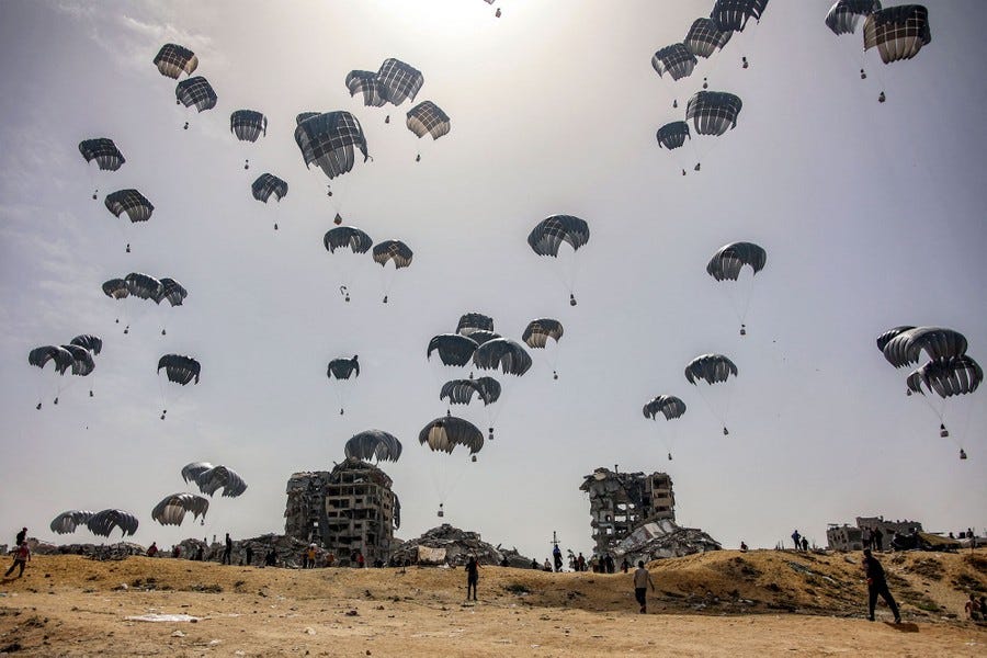 People run over low hills toward ruined buildings beneath dozens of parachutes falling from the sky, dropping aid packages.