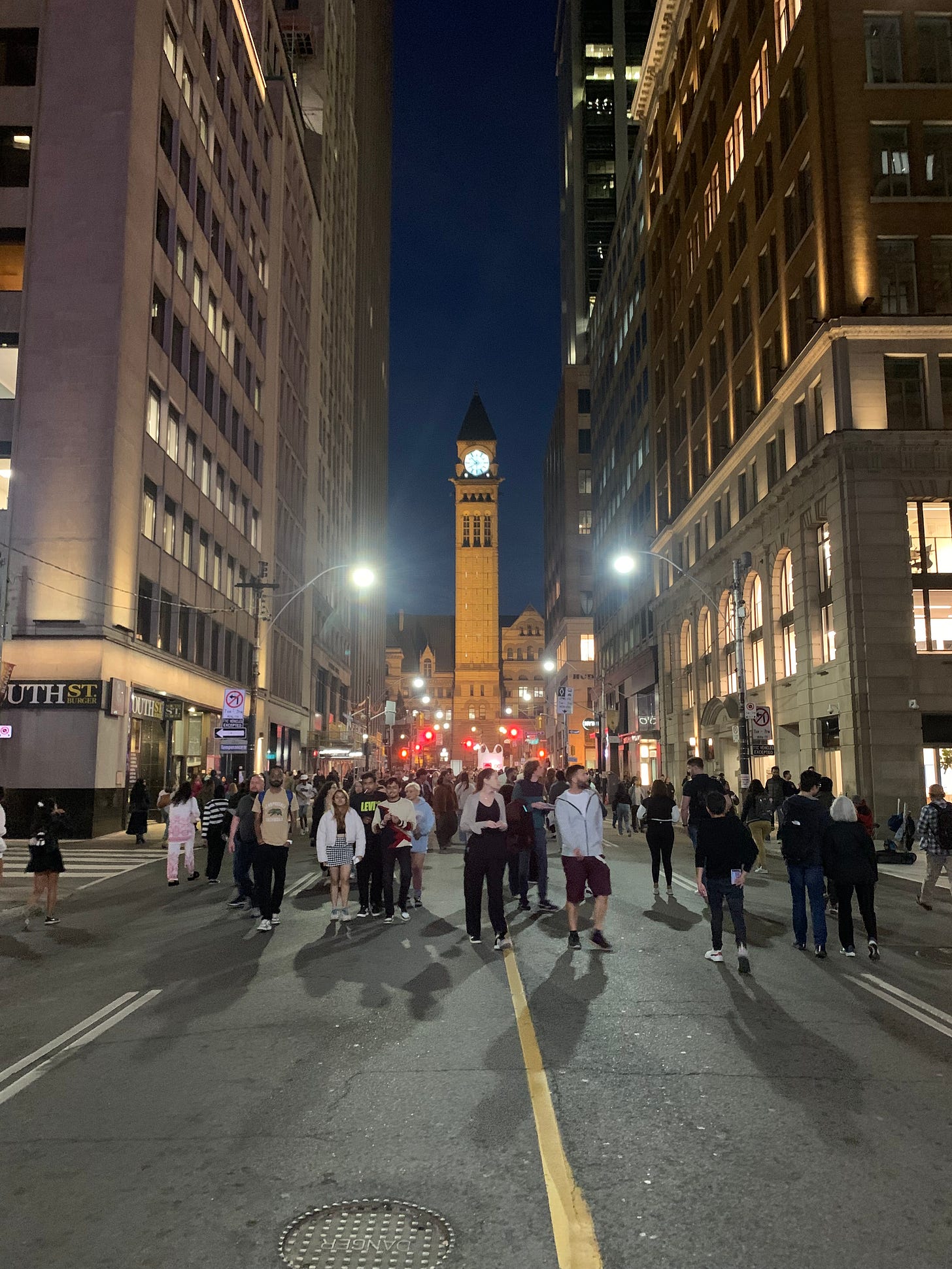 A long street with a clock tower glowing in the background at the end, and a crowd walking in the street.