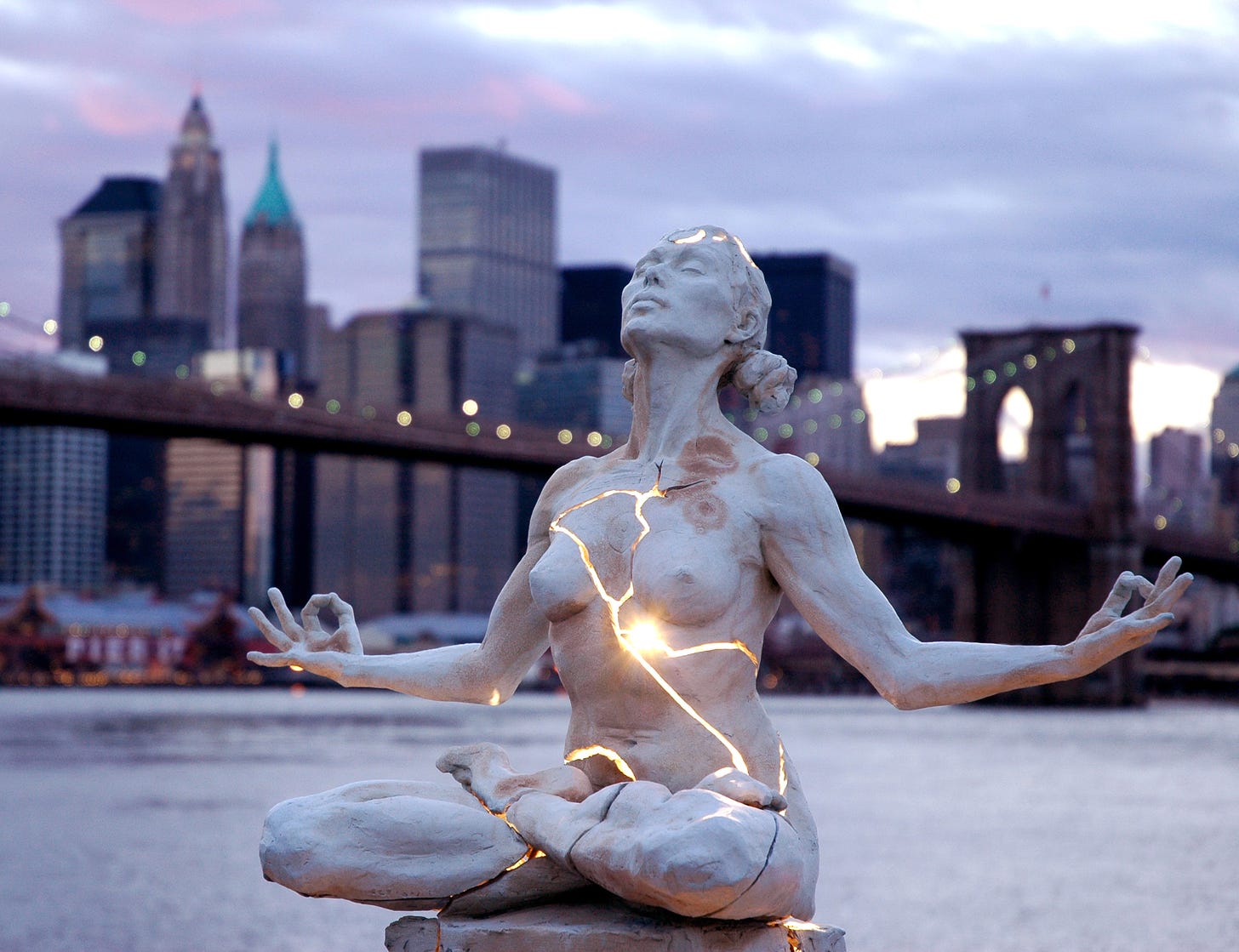 A sculpture of a woman sitting in lotus position, cracks from head to sternum to folded legs, light streaming through. In the background, the NYC skyline. Artist, Paige Bradley.