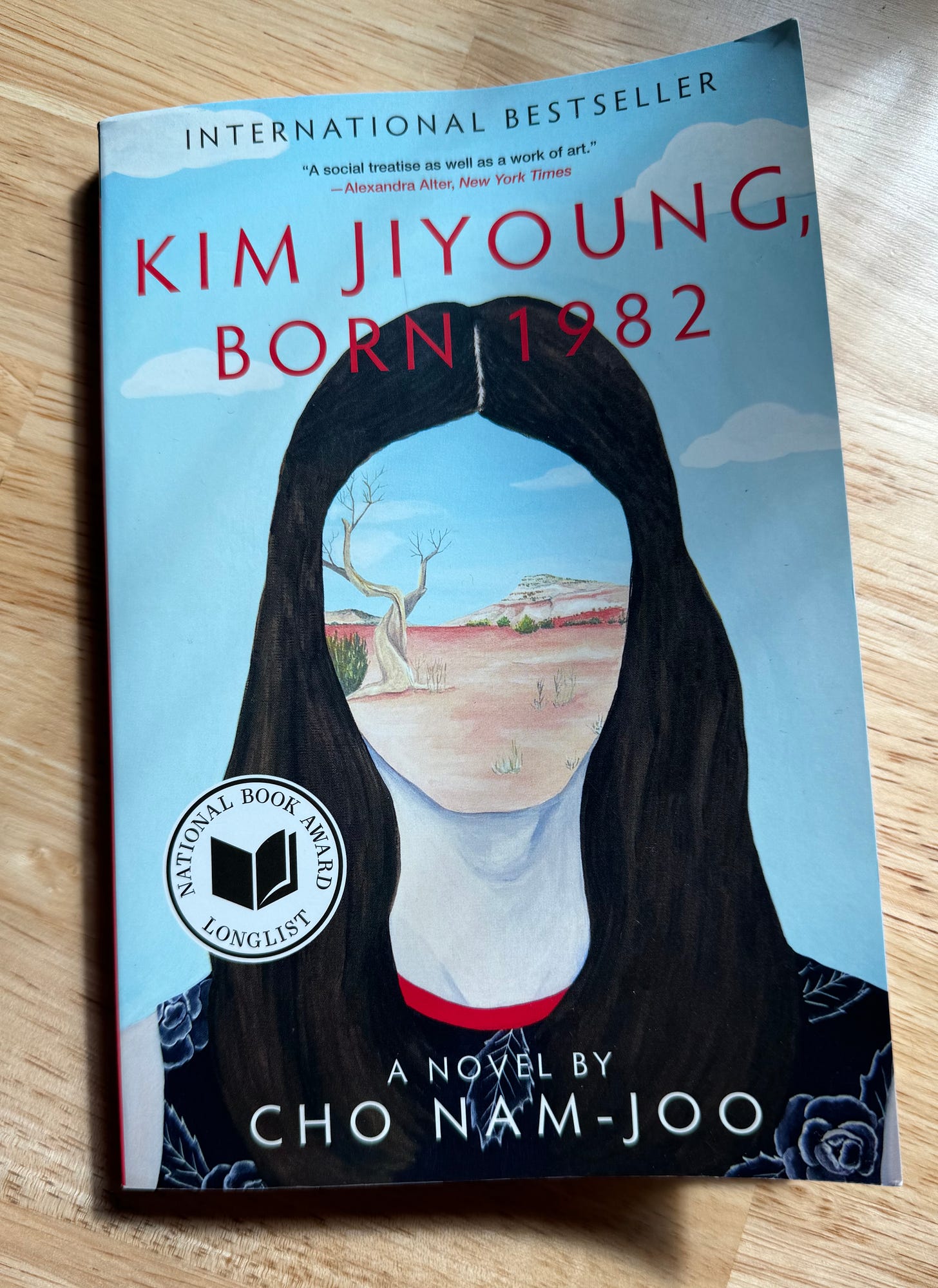 book cover of Kim Jiyoung, Born 1982 by Cho Nam-Joo. National Book Award sticker. International Bestseller. Painting of a woman with long dark hair. Instead of a distinct facial features there is a painting of a desert. 