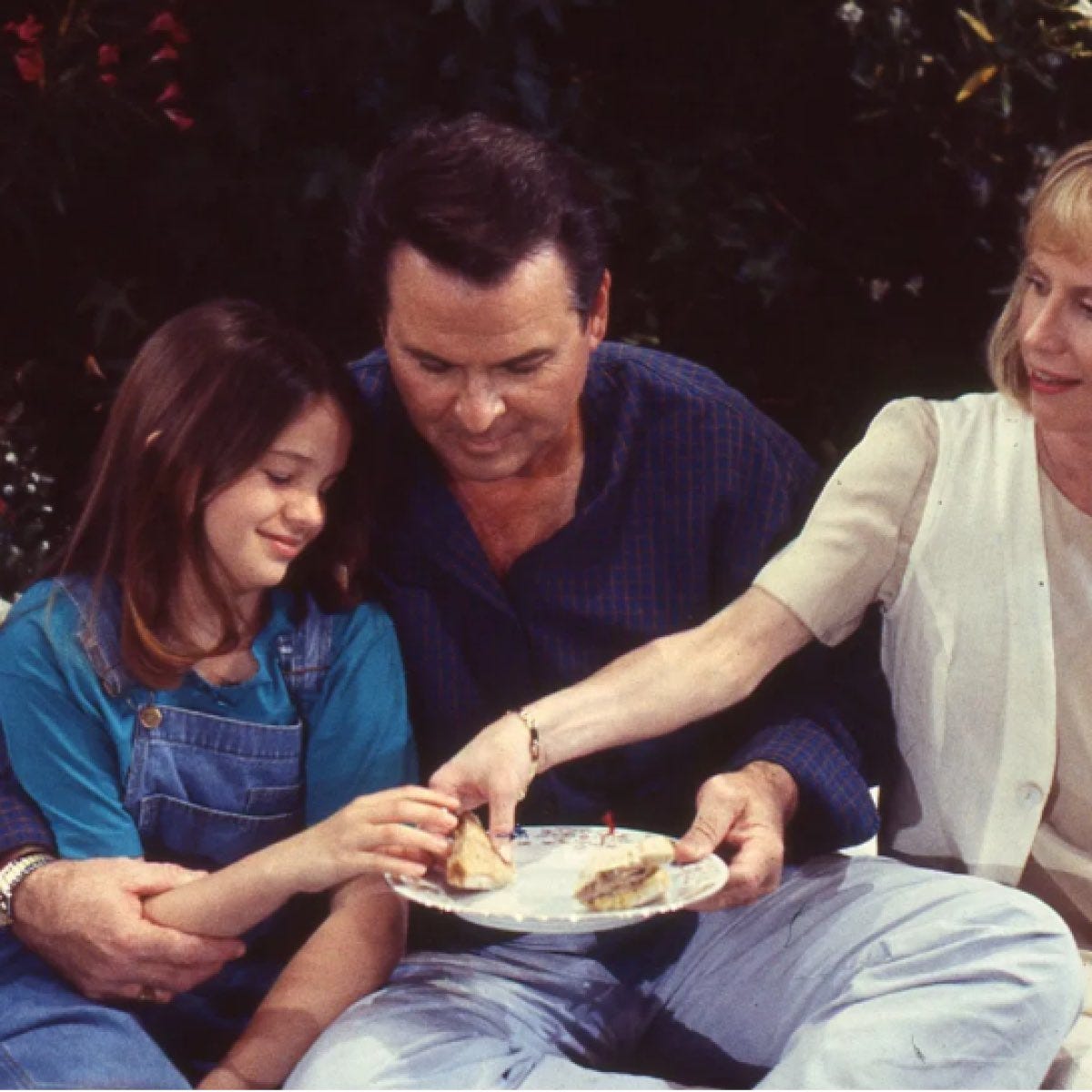 A still from "General Hospital." A very young Emily Quartermaine (Amber Tamblyn), Dr. Alan Quartermaine (Stuart Damon), and Dr. Monica Quartermaine (Leslie Charleson) sit outside sharing a sandwich together.