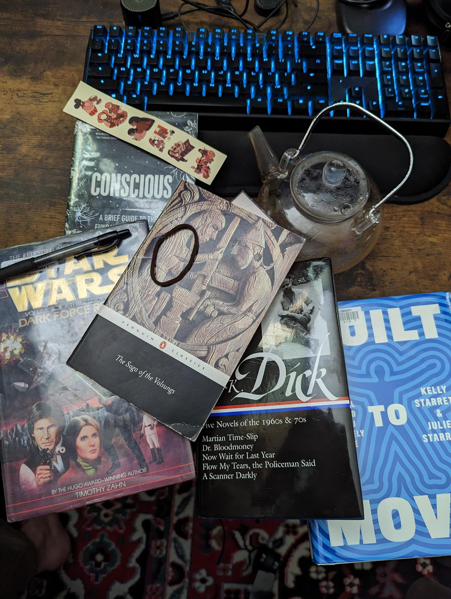 An arranged pile of books on a dark wood background, next to a clear glass teapot. It feels busy yet homey. The books are: Star Wars Dark Force Rising, a collection of Phillip K Dick novels, The Saga of the Volsungs, Built to Move, and Consciousness. by Annaka Harris.