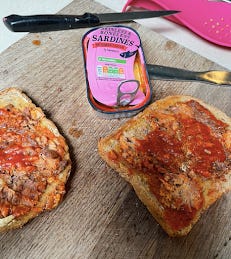 A wooden chopping board with two pieces of toast. The toast is covered with red/brown mashed up sardines. Also on the board are a used knife and an empty tin. The tin is pink with 'Skinless and boneless sardines' written on it.