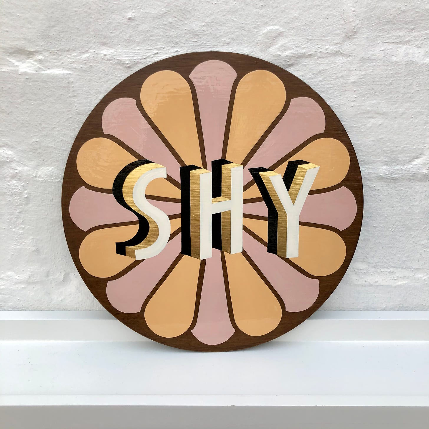 a handpainted circular sign with pink and orange petal shapes, as well as a black gold and white shadowworked lettering reading SHY. Its propped up against a white brick wall