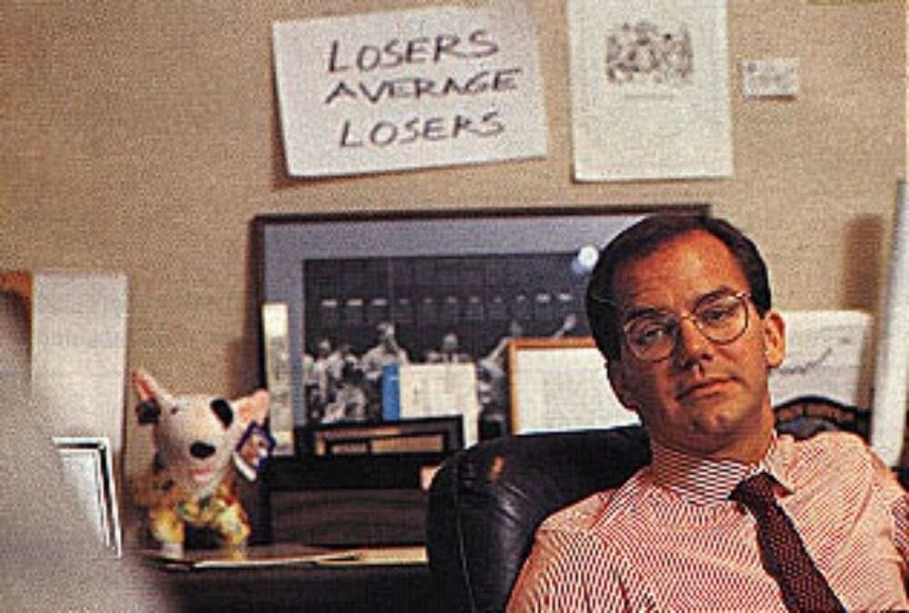 A 80's ass photo of a man staring at the camera, with the handwritten sign "LOSERS AVERAGE LOSERS" taped to the wall behind him