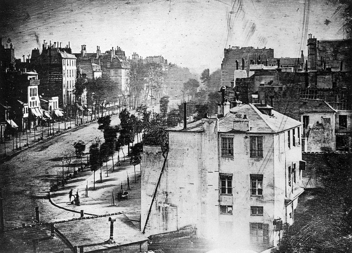 A black and white photo of a seemingly abandoned Paris street in 1838. In the bottom left corner, a blurry figure stands and gets his shoes polished.