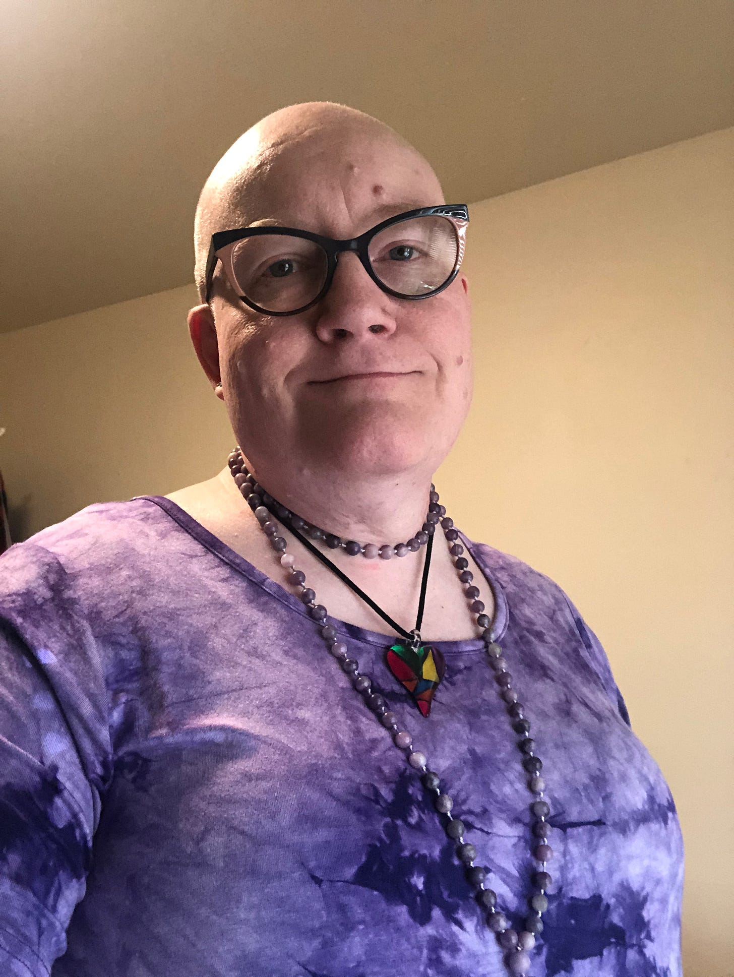 Selfie of Qid Love, a transfemme person with a bald head, wearing horn-rimmed glasses, a light purple mala, a blown glass heart pendant, and a purple tie-dyed tunic.