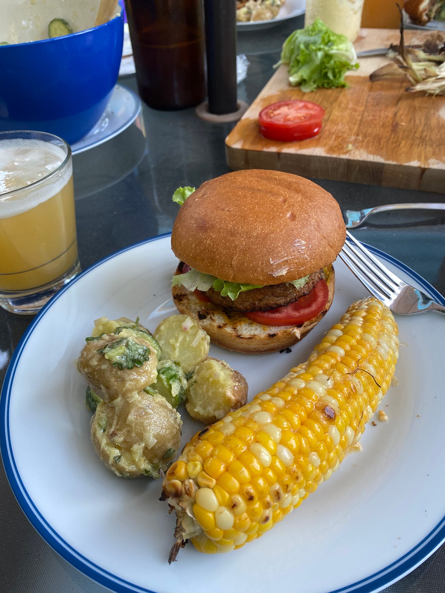 A white plate on a busy outdoor table with dishes and a cutting board in the background. On the plate are a grilled and buttered ear of corn, a small pile of skin-on german potato salad, and a veggie burger on a brioche bun with tomato and lettuce visible. A glass of hazy beer sits next to the plate.