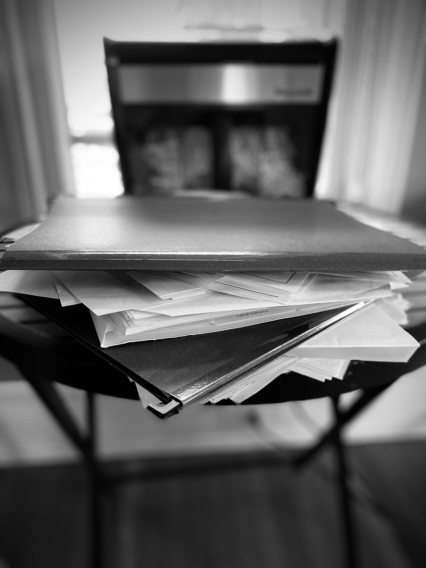 Black and white photo of a stack of files on a small round table, with a paper shredder in the background.