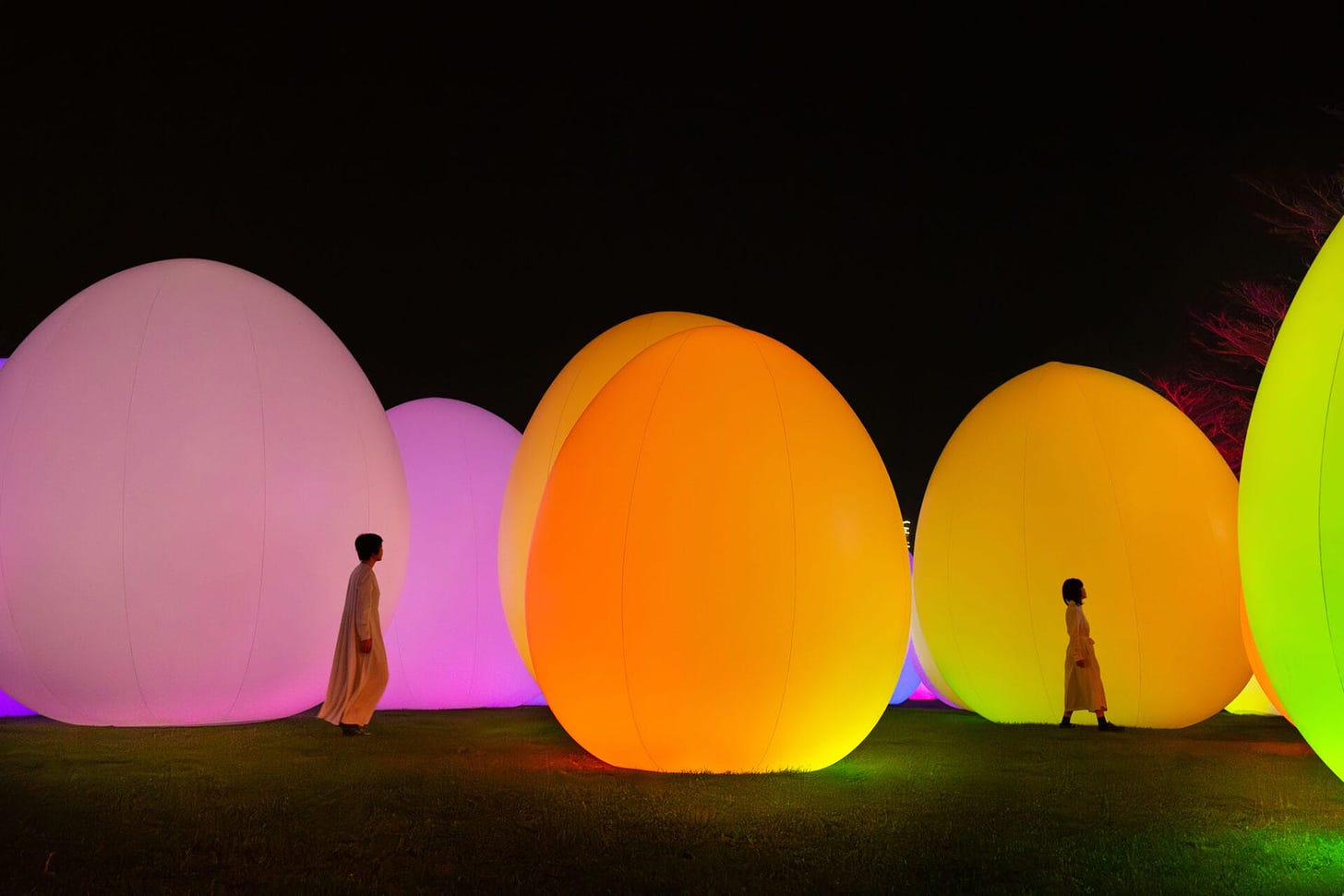 Glowing orange, pink, and light green ovoid forms. They tower in height compared to the humans standing next to them.