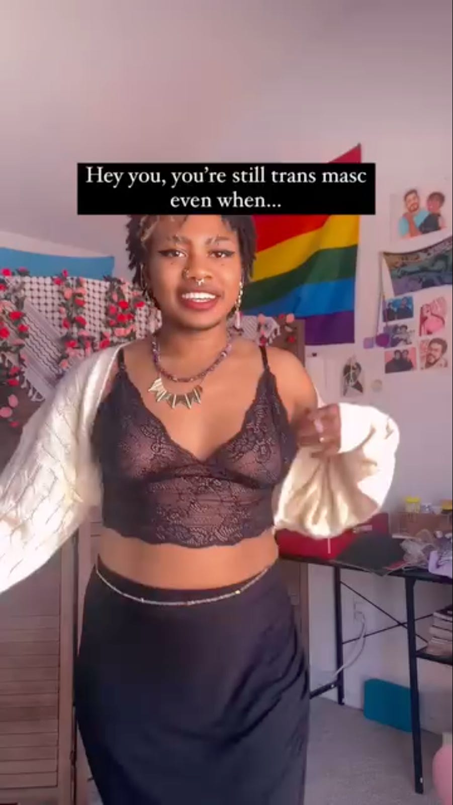 young black woman with a beard is wearing a lacy bra, a long skirt, full makeup, and jewelry. She is smiling into the camera. The text above her reads, “Hey you. You’re still transmasculine if…”