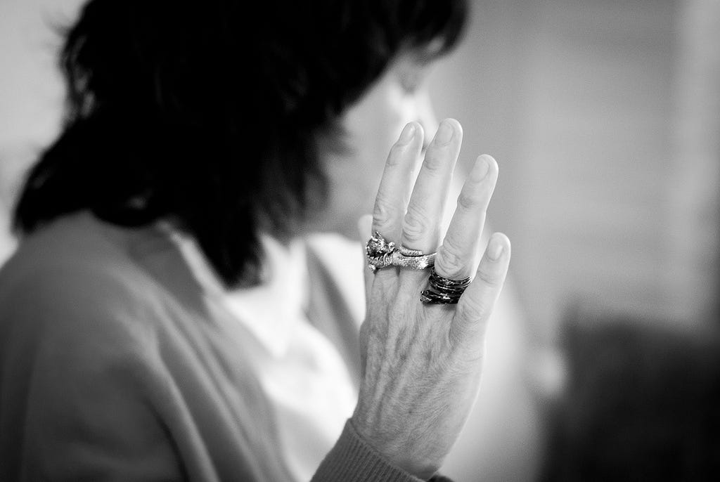 na frente, a mão de Rosa Montero, com aneis grandes nos três dedos do meio. ao fundo, o busto desfocado da escritora