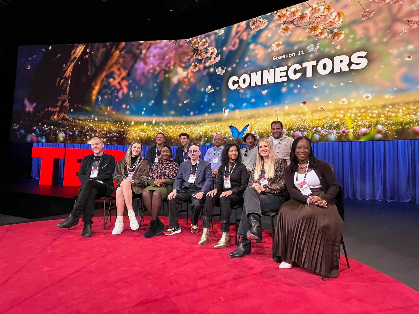 A photograph of the author and 11 other people seated on the TED stage smiling at the camera in front of a screen that reads 'Session 11: Connectors'