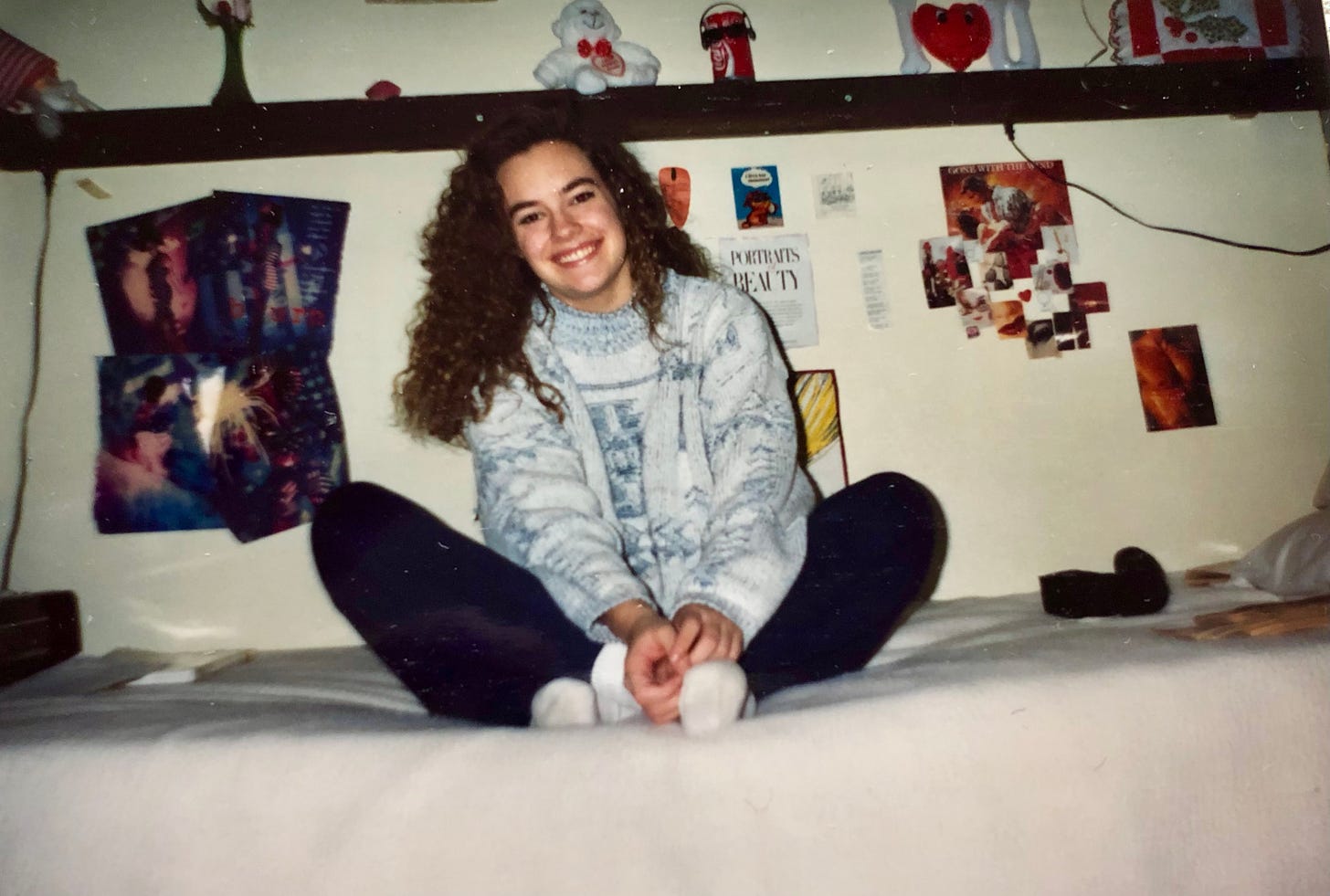 The author with long early-90s perm, sitting on her bed before a collection of wall collages and a shelf full of reminders of her childhood home. A quilted holly leaf from mom, a dancing Coke can with shades, a blow-up I love you heart...