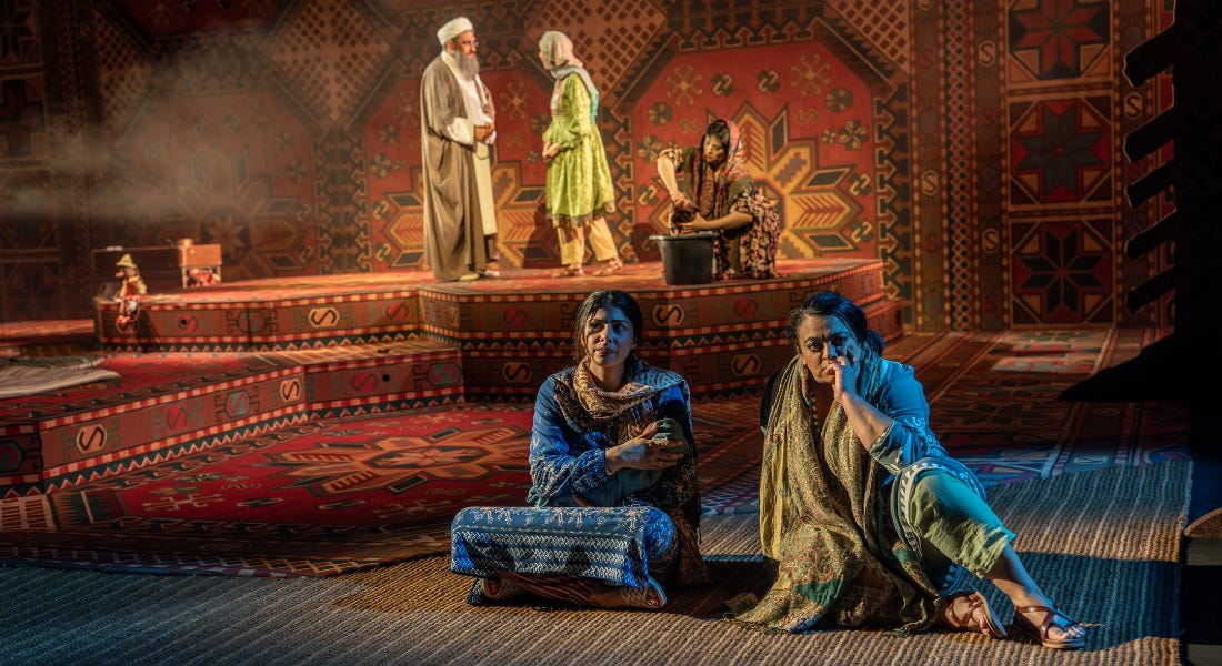 Mariam and Laila sit on a patterned rug, while a dramatic scene unfolds on stage behind them, between a young Mariam and Mullah Faizullah
