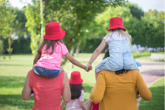 Adults and kids walking away on a park path. Two young girls ride on the shoulders of two adults, holding hands.