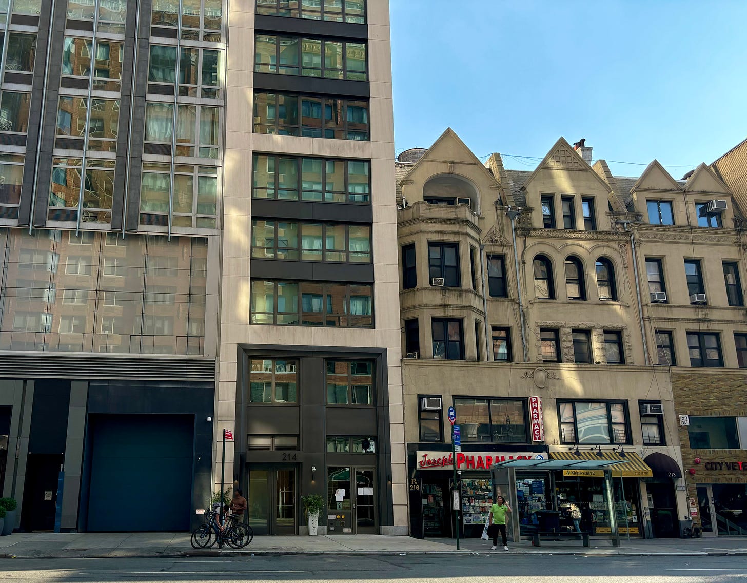 A modern apartment building of smooth stone with big windows on West 72nd, where Parker's childhood home used to be. One the left is an even larger, more modern building. On the right is a lower, old fashioned building with ornate detailing and a pharmacy in the ground floor storefront.