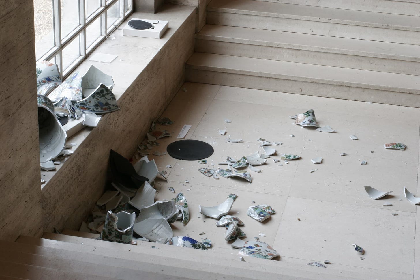 A photo of a marble stair case landing. Broken pieces of decorative vases are on a windowill and across the floor. 