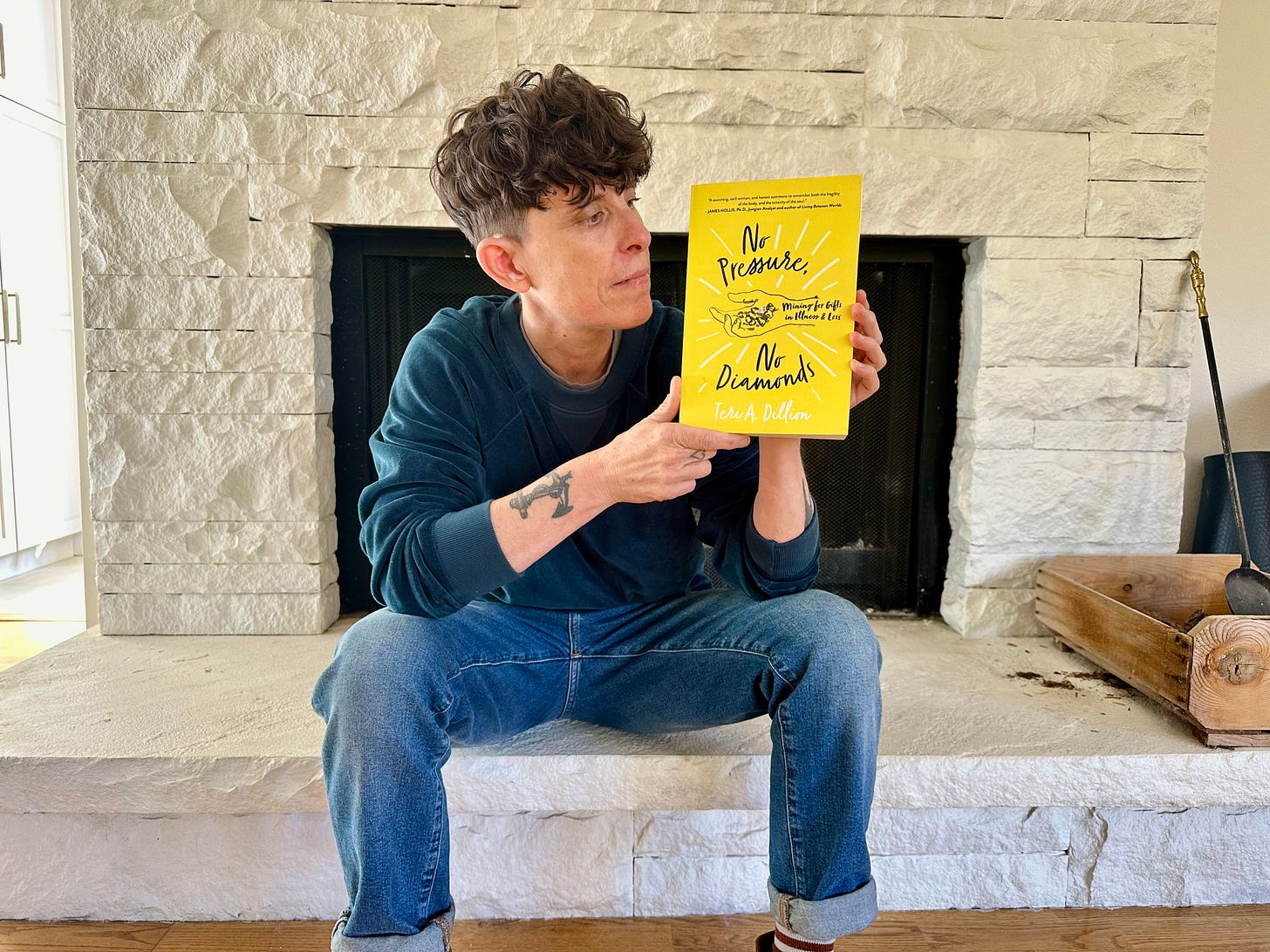 Andrea Gibson sitting in front of a white brick fireplace wearing blue jeans and a sea blue long sleeve shirt, holding the book “No Pressure, No Diamonds’ by Teri A. Dillon. 