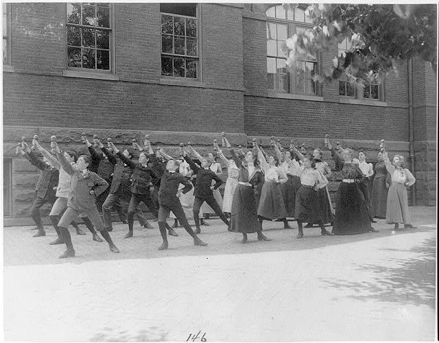 A black-and-white photo of schoolchildren and teachers standing in formation, each holding aloft a small dumbbell