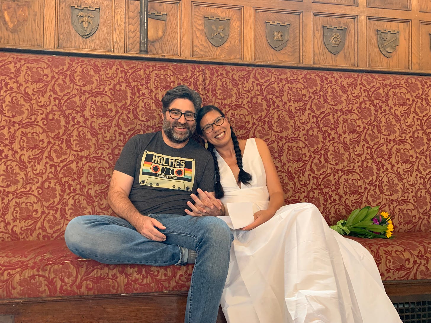 Chris and Pam sit together on an ornate bench against a wall. They lean into eachother and hold hands. They're smiling. Chris is a white man with a salt and pepper short hair and a beard and glasses. Pam is a Filipina American woman with long dark hair in braids.