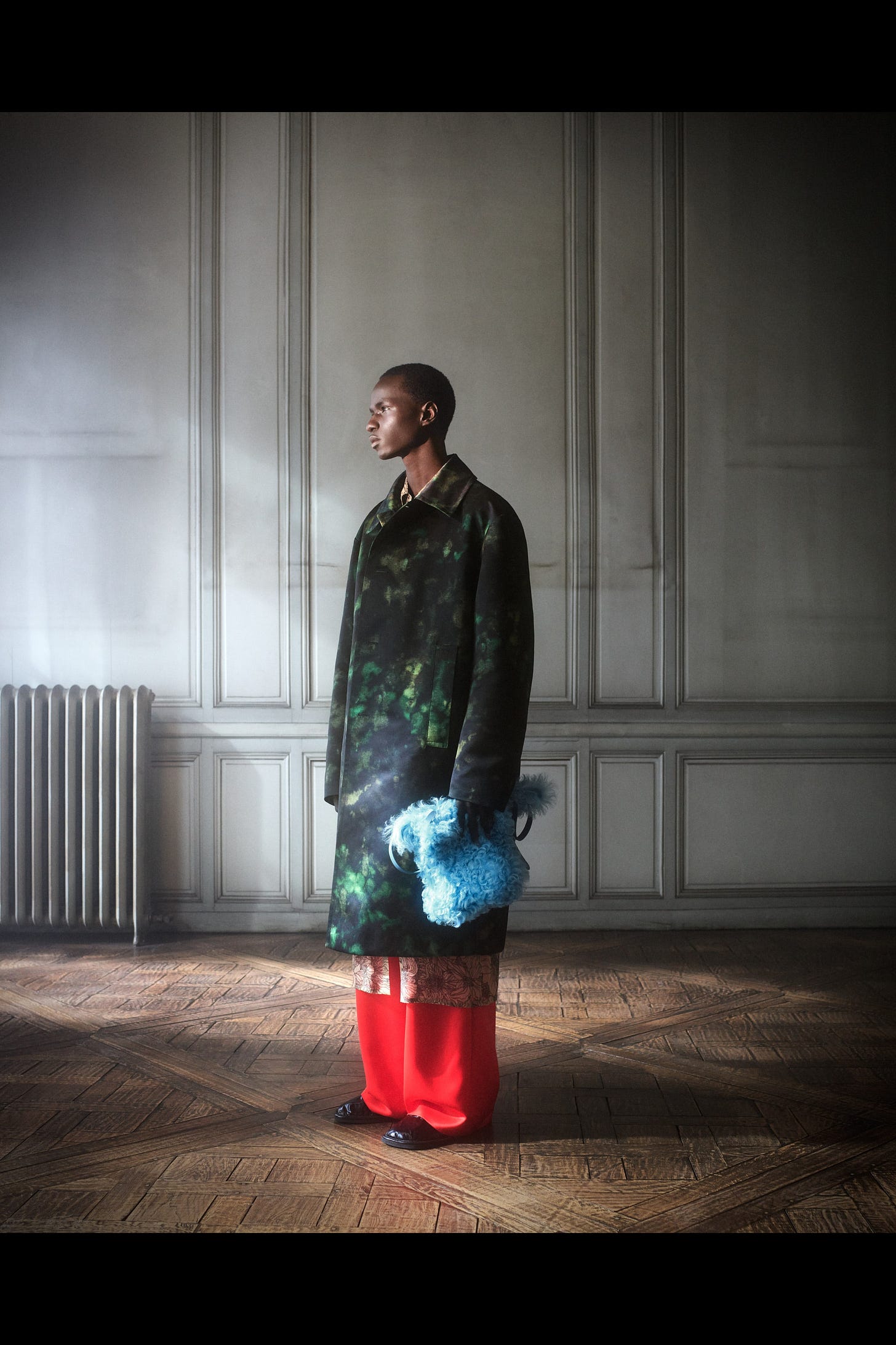 Black male standing in a grand room look left in profile wearing a dark green patterned coat and bright wide legged red trousers.