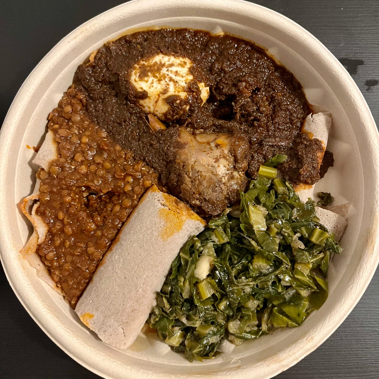 A bowl of rolled up injera, red lentils, green collard greens, and a brown chicken stew.