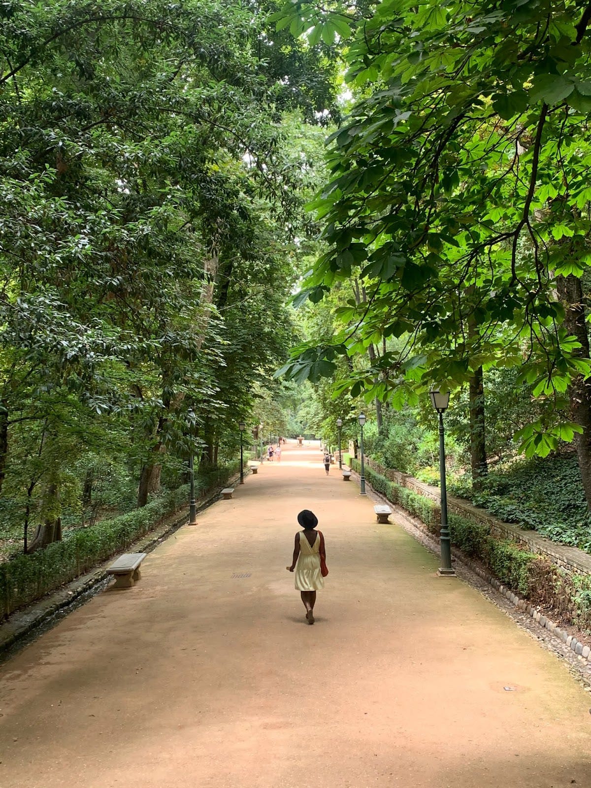 A photo of a women in a yellow dress walking through a paved path with trees