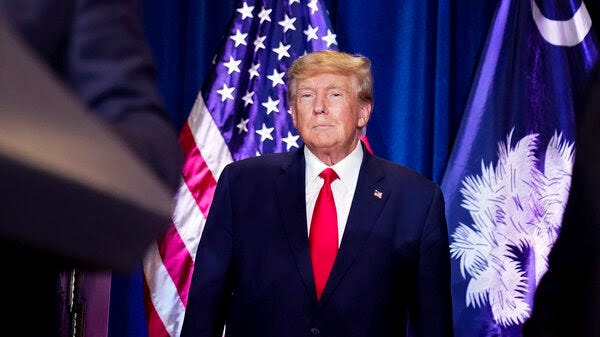 A photograph depicts former President Donald Trump in a suit and red tie, standing in front of a blue curtain and an American flag on Jan. 28 in Columbia, S.C.