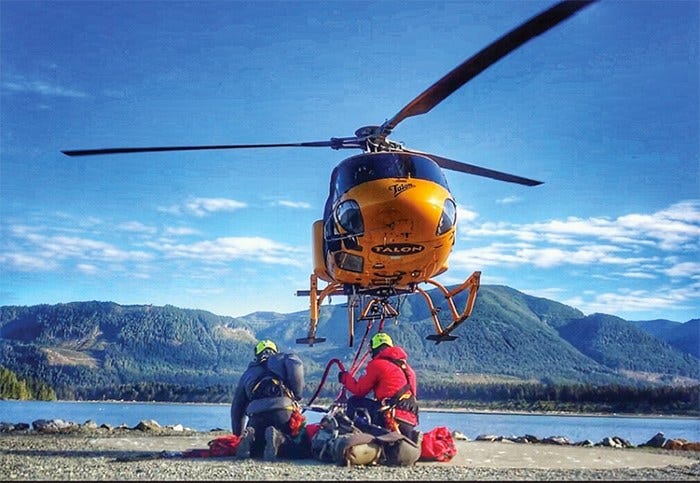 A helicopter hovers above two people in helmets with outdoors gear against a backdrop of mountains.