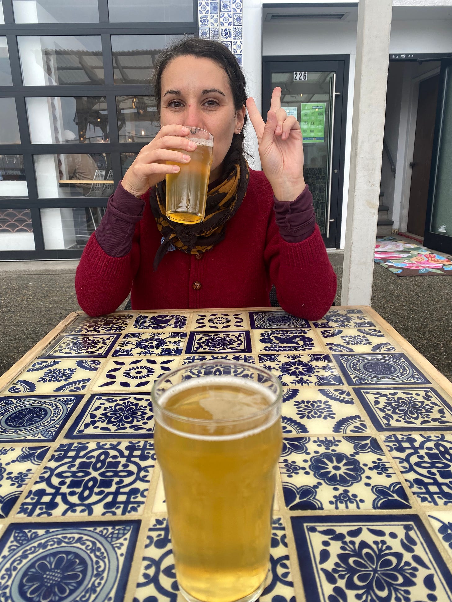 Amanda wearing a red sweater and dark patterned scarf over a maroon long-sleeved shirt, holding a beer up to her mouth and giving a peace sign. She's across a table patterned with blue and white painted tiles. My own beers is in the centre of frame in the foreground. The sidewalk in the background is wet from rain, and the garage door of the brewery is closed.