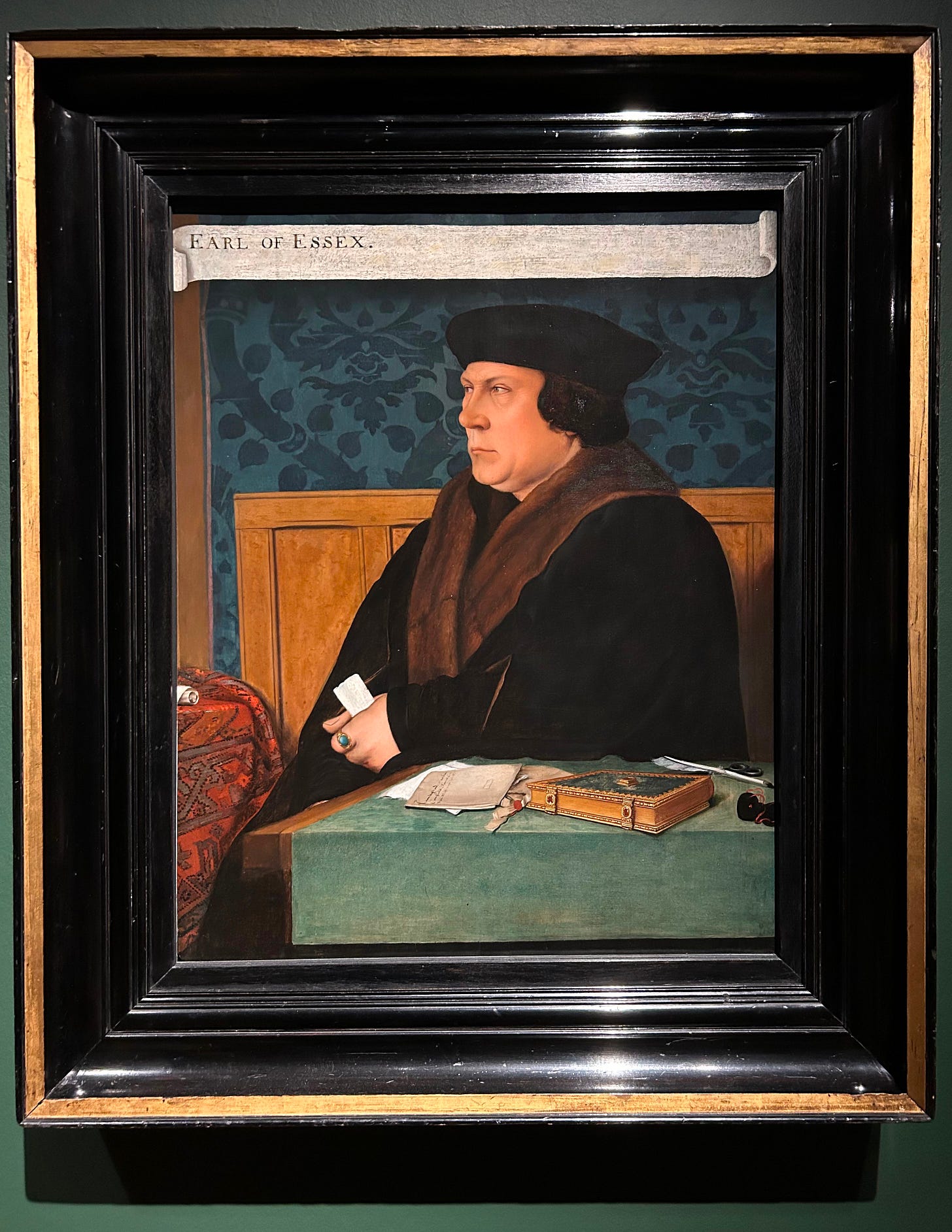 A portrait of Thomas Cromwell: a white man, not particularly handsome. He wears a black cap, and black coat with brown fur trim. In his hand - on which a turquoise ring is visible - he clutches a roll of paper. On his desk he has a book, a seal pouch, a quill and ink, and letters. There is a legend at the top of the paining which reads “Earl of Essex”.