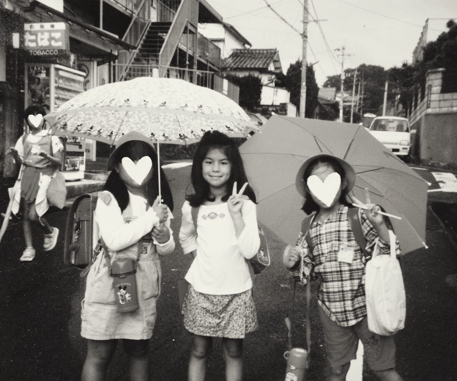 The author, Erika, is 7 years old and standing in between two Japanese girls from her class. Erika is posed making a peace sign and each friend is carrying an umbrella. They are in the middle of their school walking route.