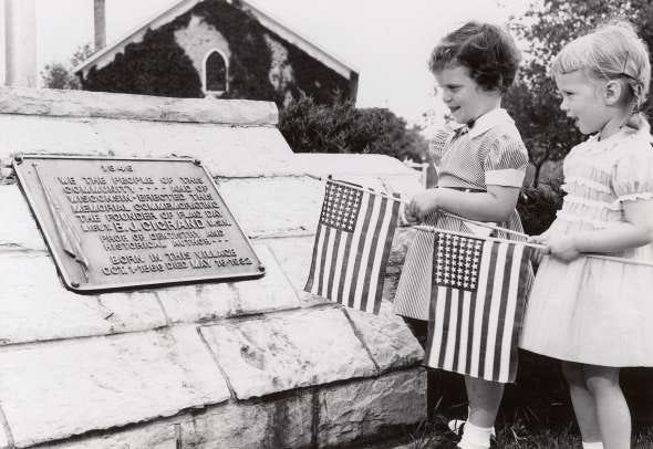 Two young girls hold American flags next to a plaque honoring Bernard J. Cigrand in Waubeka, Wisconsin, around 1949 as part of a Flag Day commemoration. UW Digital Collections